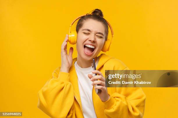 Trendy girl singing favorite song out loud in phone as mic, wearing wireless headphones, isolated on yellow background. Karaoke online app.