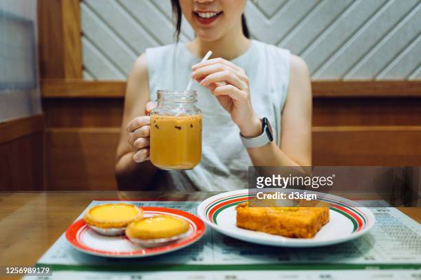 young asian woman enjoying freshly baked hong kong style egg tart and french toast with iced milk tea in a local tea restaurant - egg tart stockfoto's en -beelden