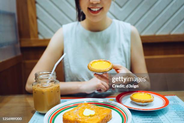 young asian woman enjoying freshly baked hong kong style egg tart and french toast with iced milk tea in a local tea restaurant - egg tart stockfoto's en -beelden