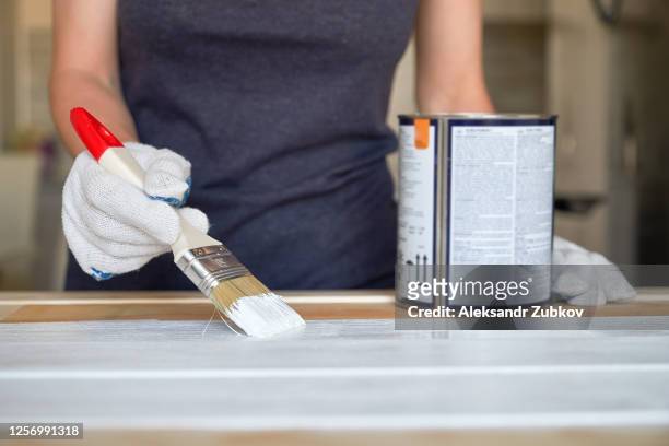 brush with white paint in the hands of a woman in protective gloves. next to it is a can of paint. painting and repair work. a female painter paints boards and surfaces, and makes a wooden photophone herself. - emaille stock-fotos und bilder