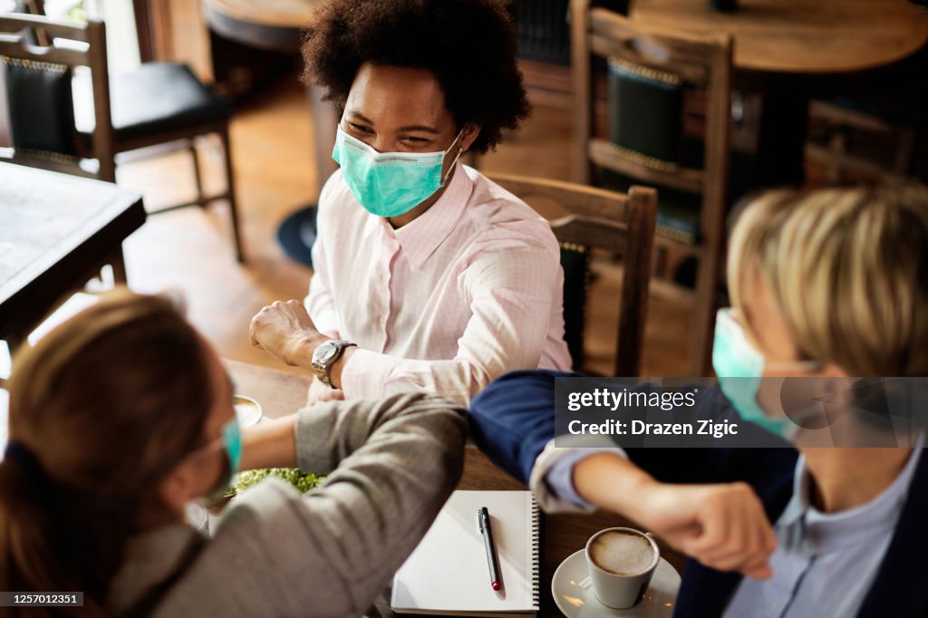 Happy businesswomen with face masks elbow bumping in a cafe.