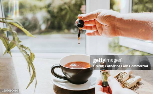 hand dropping cbd oil into a cup of tea, surrounded by cannabis plants - cannabidiol stock pictures, royalty-free photos & images