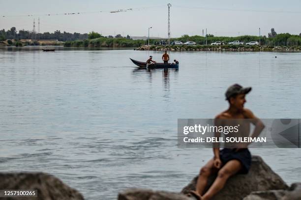 Youth sits on a rock as others row their boat in the Euphrates river in the northern Syrian city of Raqa on May 19, 2023.