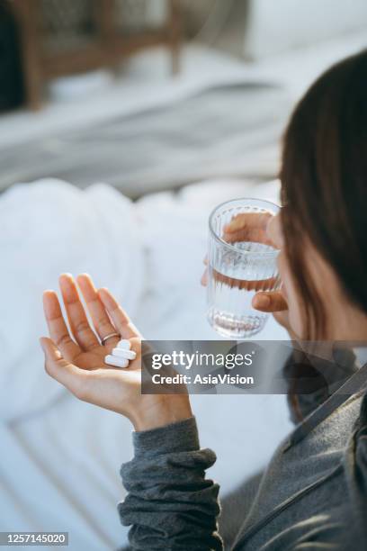 ベッドに横たわって気分が悪く、コップ一杯の水で薬を手に取る若いアジア人女性のクロップドショット - glass of water hand ストックフォトと画像