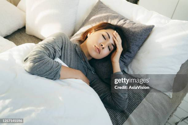 young asian woman with hand on forehead lying in bed and feeling sick - heat illness stock pictures, royalty-free photos & images