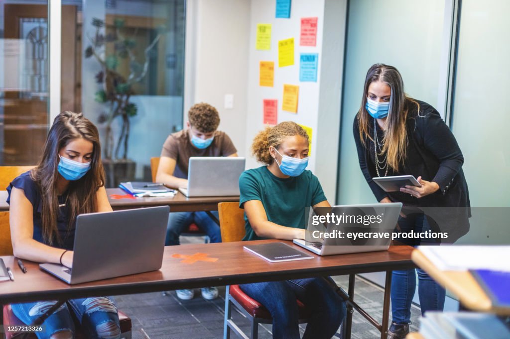 Gymnasiasten und Lehrer tragen Gesichtsmasken und soziale Abstellzeit im Klassenzimmer, die an Laptop-Technologie arbeiten