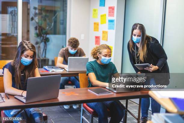 high school students and teacher wearing face masks and social distancing in classroom setting working on laptop technology - high school stock pictures, royalty-free photos & images