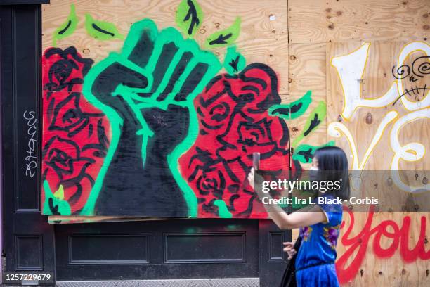July 18: A bystander wearing a mask stands in front of a painting on a boarded up storefront with the black power fist and roses holds up her phone...