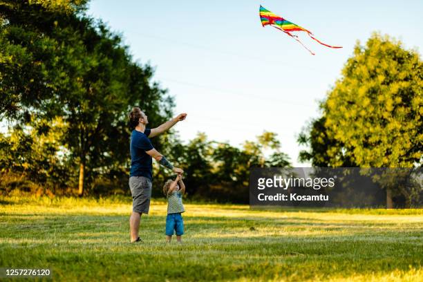 modern father teaching his toddler boy how to fly a rainbow kite on a summer day - white dragon tattoo stock pictures, royalty-free photos & images