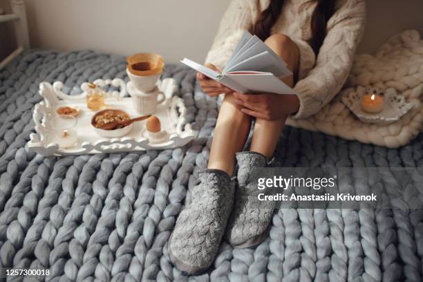 woman with long hair drinking hot coffee and reading book in bed. woman in woolen socks and sweater sitting on wool chunky merino plaid. cozy winter morning concept. - cosy stock-fotos und bilder