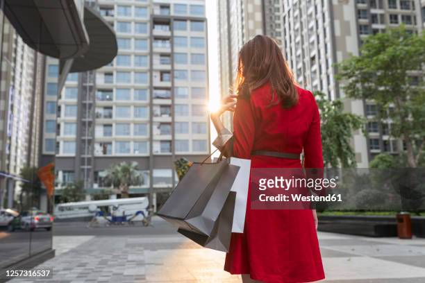 young woman carrying shopping bags walking on city street - red dress stock-fotos und bilder