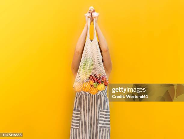 young woman holding fruits in mesh bag while standing against yellow background - reusable bag isolated stock pictures, royalty-free photos & images