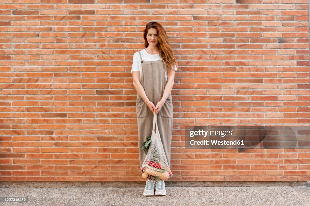 Young woman with long hair holding groceries in mesh bag while standing against brick wall