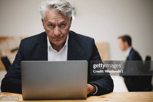 senior businessman working on laptop, sitting in office with collueague - suspicion stock-fotos und bilder