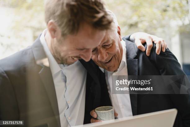 father leaning on shoulder of his son, looking at laptop, smiling - senior adult stock-fotos und bilder