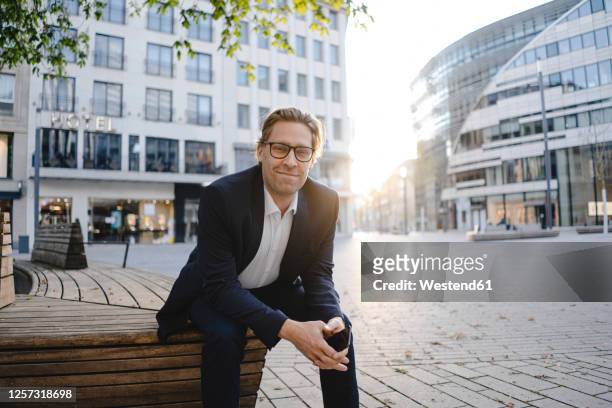 portrait of smiling businessman sitting on a bench in the city - veste homme photos et images de collection