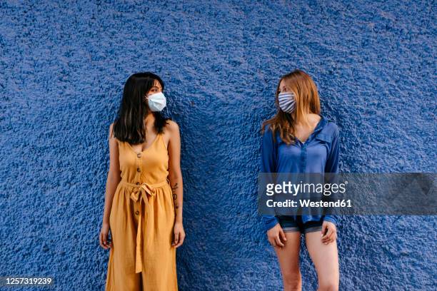 female friends wearing masks looking at each other while maintaining distance against blue wall in city - short hair photos et images de collection