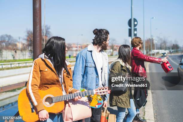 young man hitchhiking while standing with friends on street in city during sunny day - hitchhike stock-fotos und bilder