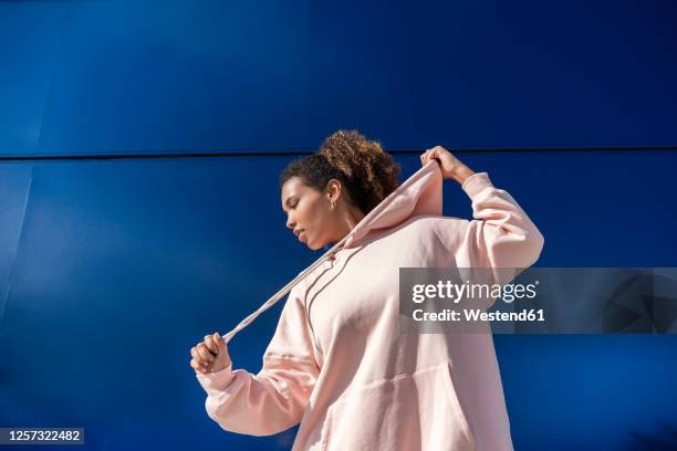 smiling young woman wearing pink hoodie sweater in front of blue wall - kapuzenoberteil stock-fotos und bilder