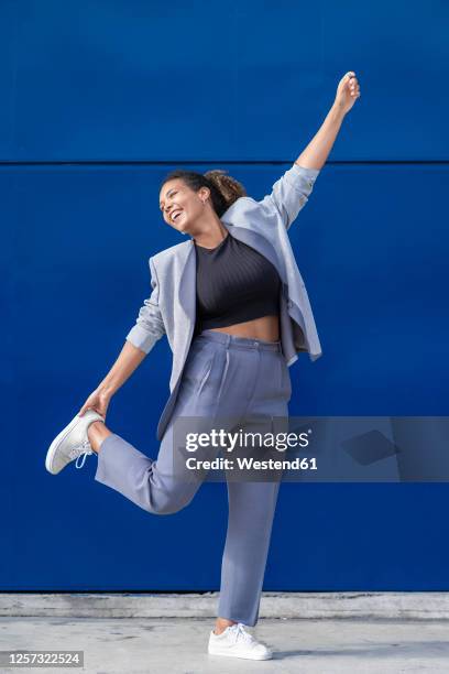 smiling businesswoman with raised arm in front of blue wall - blazer foto e immagini stock