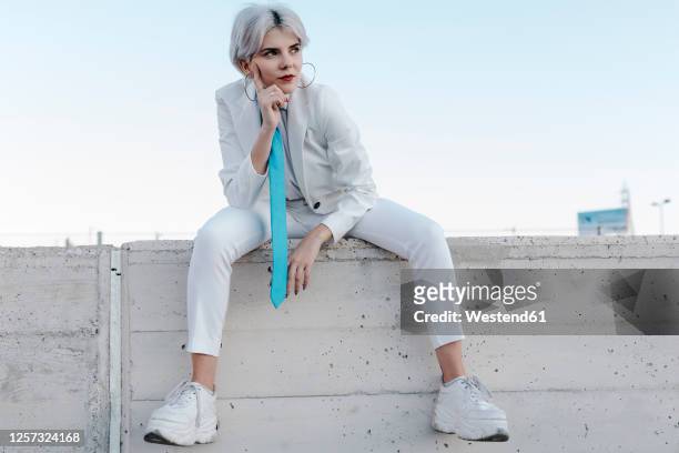 thoughtful young woman wearing white suit sitting on retaining wall against clear sky - capelli grigi foto e immagini stock