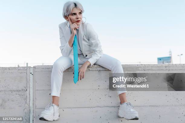 thoughtful young woman wearing white suit sitting on retaining wall against clear sky - woman silver hair young ストックフォトと画像