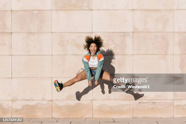 excited young woman screaming while jumping against wall during sunny day - saltare foto e immagini stock