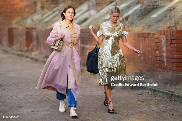 Alexa Chung wears a pale pink transparent trench coat with a beige part, a belt, a bag, blue jeans, white sneakers ; a guest wears a golden sequined...