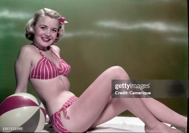 Young blonde woman in red stripped two-piece bathing suit sitting on white towel with beach ball.