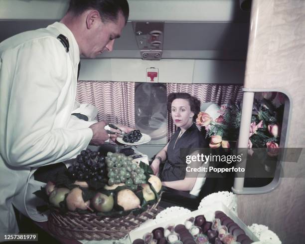 Female passenger is served a plate of grapes from a large basket of fruit by an air steward in the passenger cabin of a Bristol Britannia medium to...