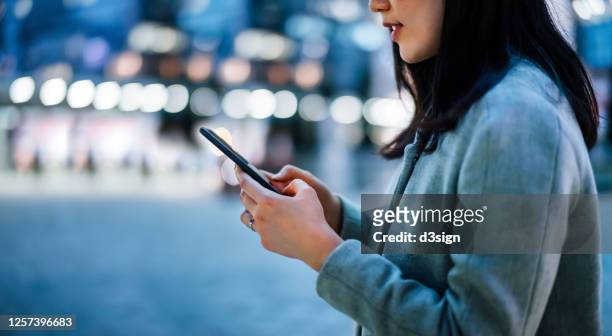 close up of young asian businesswoman using smartphone while commuting in downtown city street, with illuminated city street lights in background - applicazione mobile foto e immagini stock