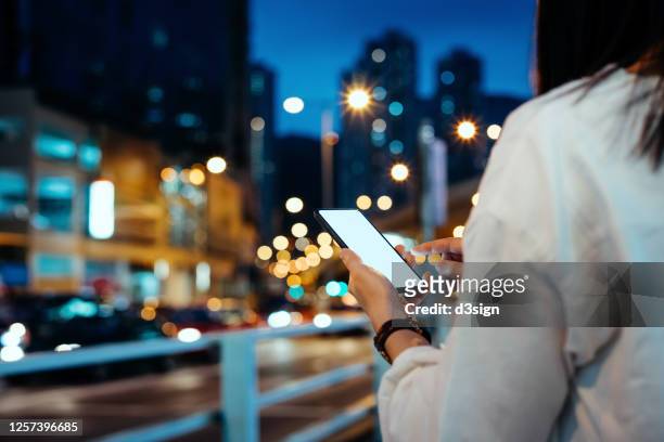 young asian woman requesting a taxi ride with mobile app device on smartphone in downtown city street with illuminated street lights and busy traffic after work - sharing economy stock pictures, royalty-free photos & images