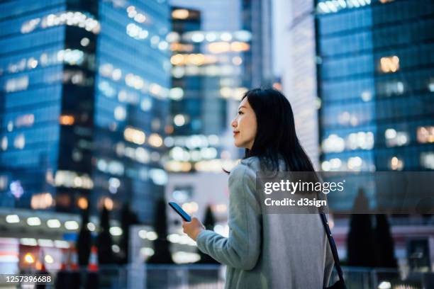 successful young asian businesswoman using smartphone on the go in financial district after work, against urban city scene with illuminated corporate skyscrapers - china banking regulatory commission stockfoto's en -beelden