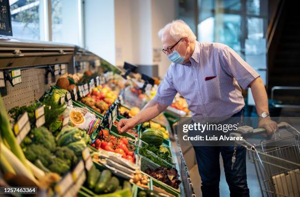 senior man with face mask buying vegetables in grocery store - face mask coronavirus stock-fotos und bilder