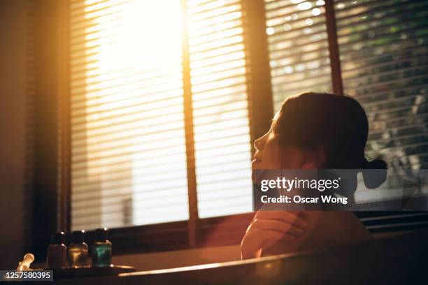 staycation: young woman relaxes in bath and has spa at home with her eyes closed against sunset - japanese women bath stock pictures, royalty-free photos & images