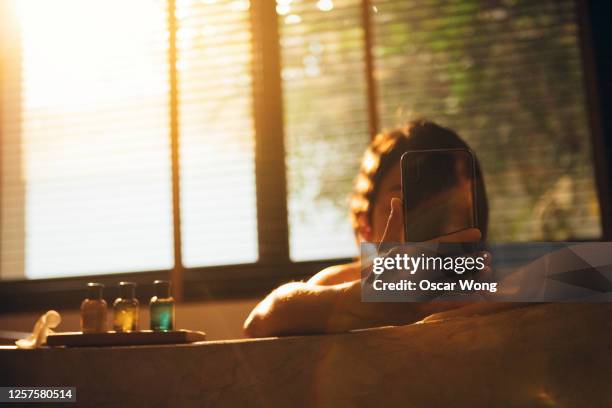 staycation: young woman checking work email on smartphone while taking a bubble bath at home with lens flare effect - bathing in sunset stockfoto's en -beelden