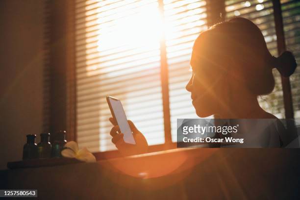 staycation: young woman using mobile phone while taking a bubble bath at home with lens flare effect - japanese women bath stock-fotos und bilder