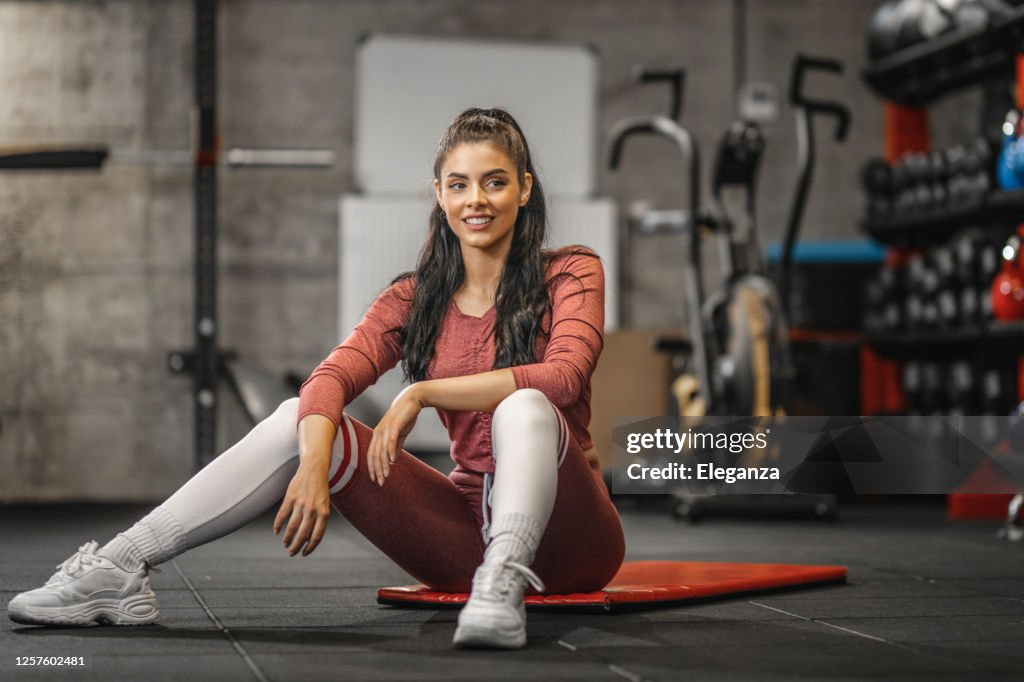 Retrato de una mujer deportiva sonriendo y sentada en la alfombra de ejercicio en el gimnasio