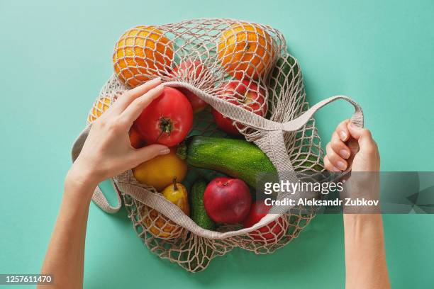 fresh juicy fruits and vegetables, products in a reusable shopping bag. a girl or woman takes or lays out products from a string bag made from recycled materials on a green pastel background. vegetarianism, veganism. no plastic. - obst stock-fotos und bilder