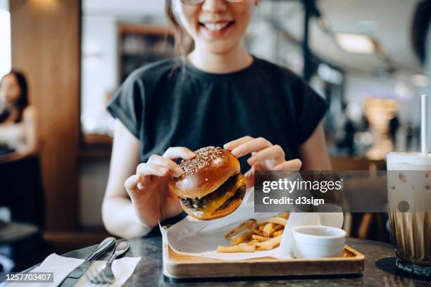 smiling young asian woman enjoying freshly made delicious burger with fries and a glass of iced coffee in a cafe - unhealthy eating ストックフォトと画像