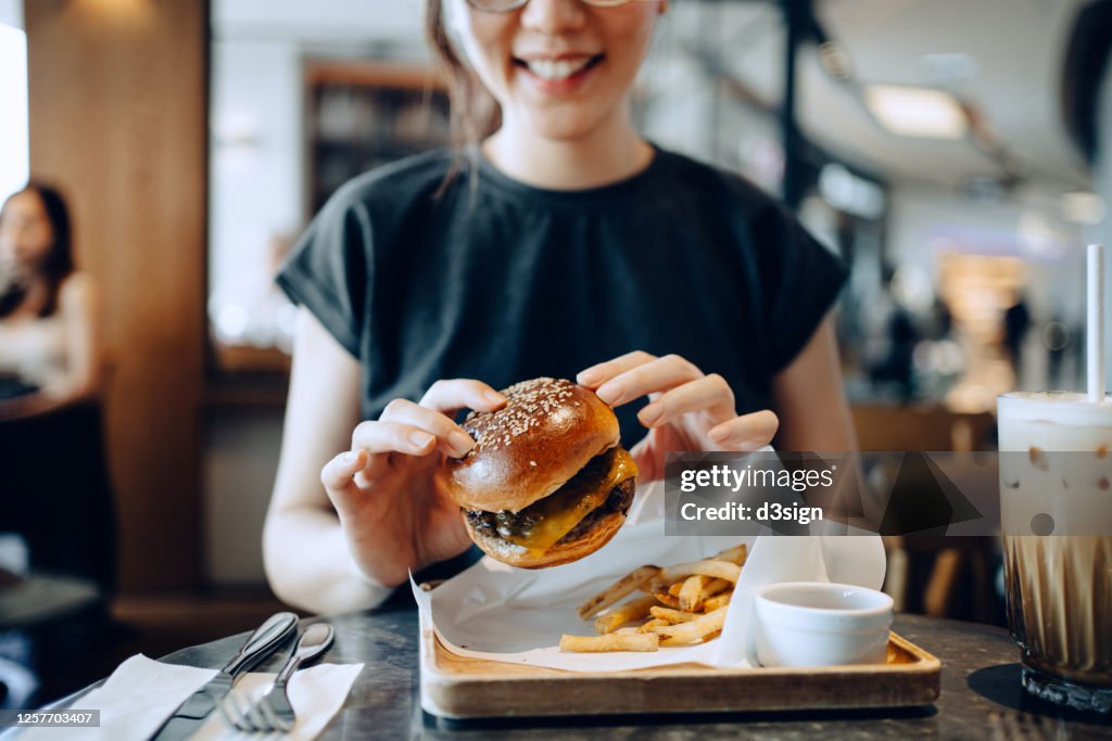 Smiling young Asian woman enjoying freshly made delicious burger with fries and a glass of iced coffee in a cafe