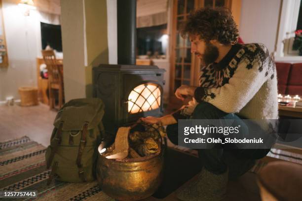 hombre sentado cerca de estufa de leña en cabaña acogedora - wood burning stove fotografías e imágenes de stock