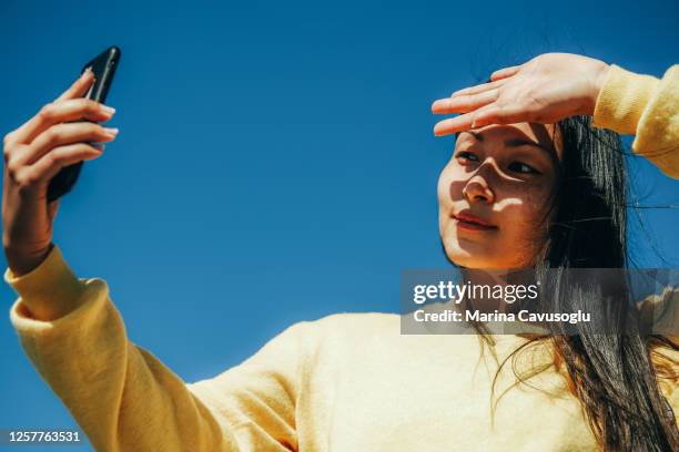 young asian girl in yellow sweater taking a selfie with her phone in a sunny day. - city life authentic stockfoto's en -beelden