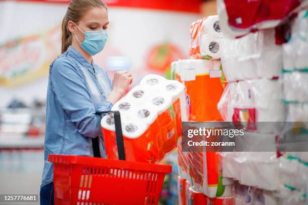 young woman wearing protective face mask putting a pack of toilet paper in shopping basket - buying toilet paper stock pictures, royalty-free photos & images