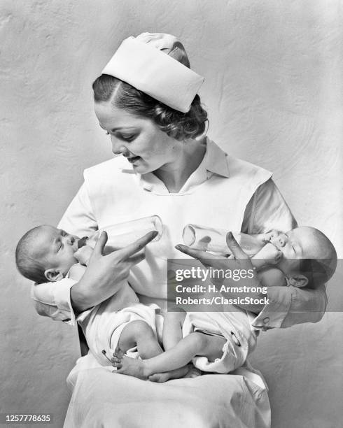 1930s Smiling Nurse Sitting Holding Juggling Two Newborn Twin Baby Girl Sisters In Her Arms Feeding Them With Bottles
