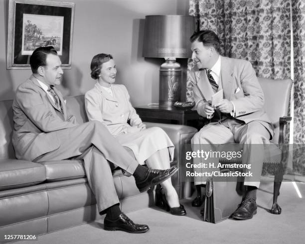 1940s Smiling Man Salesman Holding Brochure Insurance Policy Talking Leaning Toward Couple Husband And Wife Sitting On Sofa