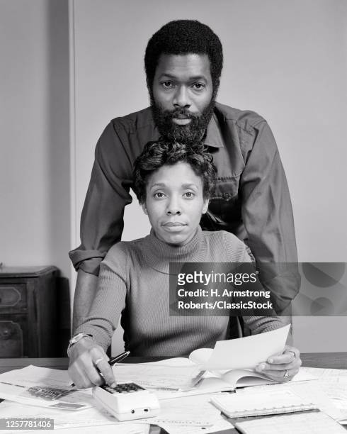 1970s African-American Couple Looking At Camera Man Standing Behind Woman Sitting At Table With Papers Bills Hand On Calculator