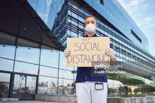 responsible doctor holding a sign with a message ''keep social distancing" during covid-19 pandemic - state of emergency sign stock pictures, royalty-free photos & images