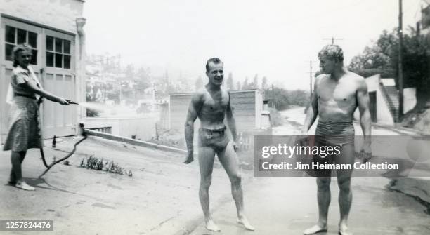 Woman aims a garden hose at two men in swimming trunks on the driveway of their Silver Lake home in Los Angeles, circa 1947.