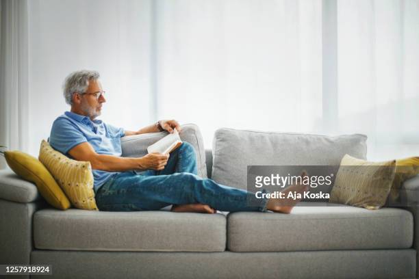 reife mann lesen ein buch auf dem sofa. - reading stock-fotos und bilder